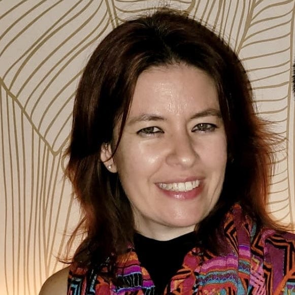 Leigh Nelson-Woman wearing a colorful patterned scarf against a decorative wall.